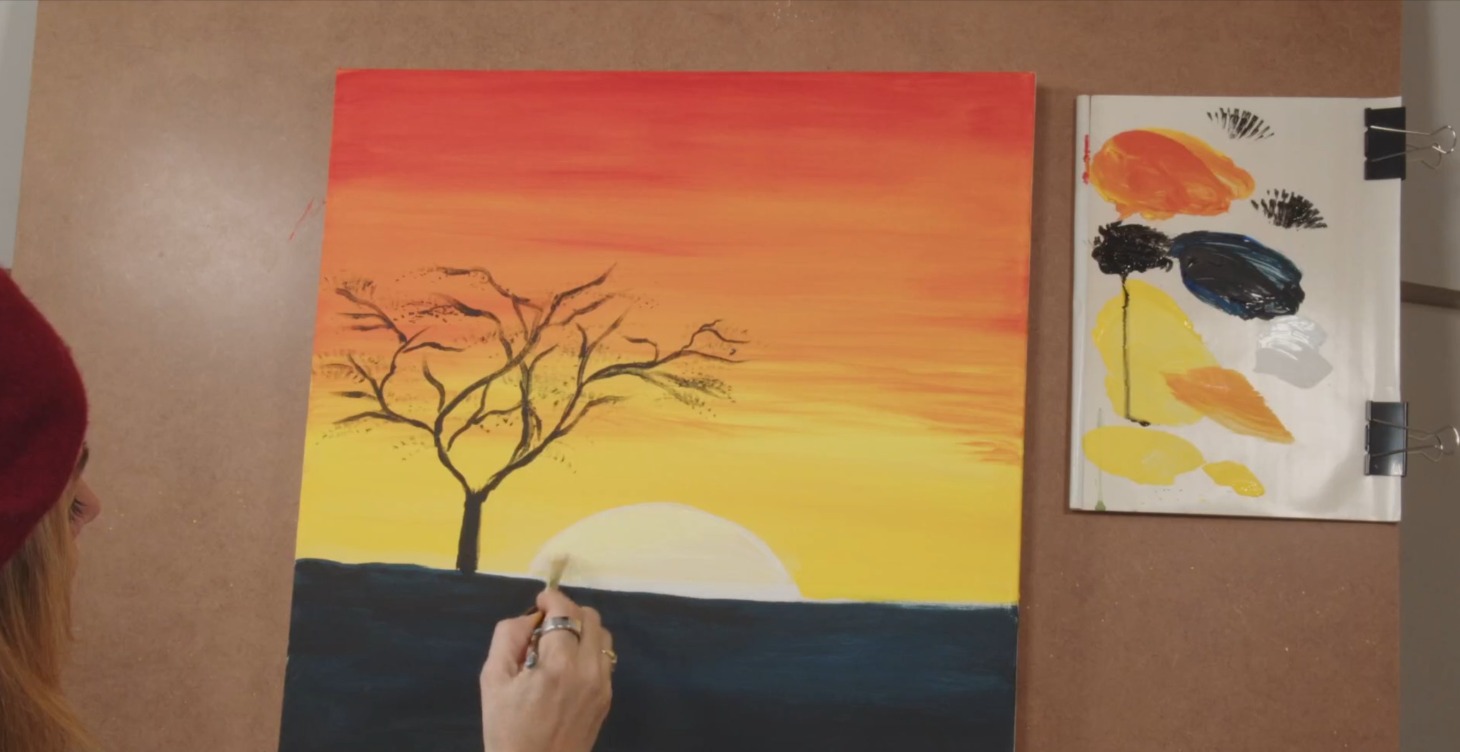 Painting of an orange sunset with bare tree in progress next to a paint palette