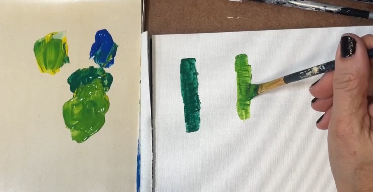 Brush testing two green color mixes on canvas next to a palette of mixed greens