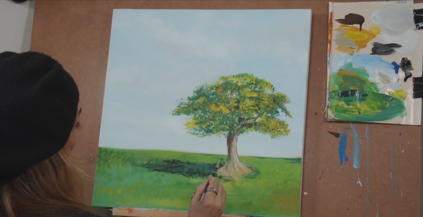 Artist painting a leafy green tree in a meadow landscape on canvas